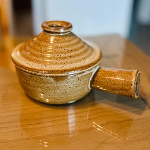 Pode incluir: Uma panela de cerâmica marrom com tampa e alça. A panela tem um esmalte salpicado e é perfeita para servir uma refeição pequena.