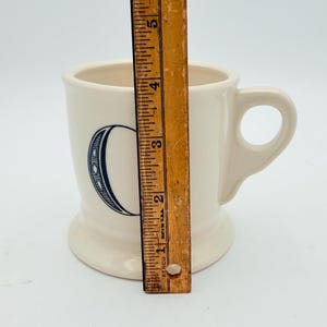 Puede incluir: Taza de cer&aacute;mica color crema con una gran letra "C" azul marino. La taza tiene un asa y una base ancha. Una regla de madera est&aacute; al lado, midiendo aproximadamente 10 cm de altura.