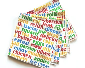 Puede incluir: Una pila de paños de cocina blancos con texto colorido. El texto incluye palabras relacionadas con la comida como "limones", "tocino" y "manzanas". Las palabras están impresas en una variedad de colores, incluyendo rojo, azul, verde y amarillo.
