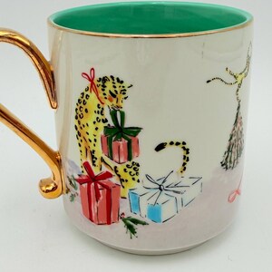 K&ouml;nnte beinhalten: Eine Keramik-Tasse mit goldenem Henkel und Rand, mit einem Leoparden-Design mit Geschenken. Der Innenraum ist gr&uuml;n. Die Geschenke sind rot, gr&uuml;n und blau. Die Tasse hat ein festliches Urlaubs-Thema.