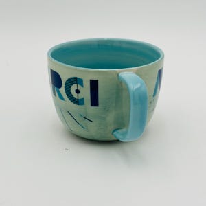 May include: A light blue ceramic mug with a white and blue geometric design. The mug has the word "Merci" written on it in blue.