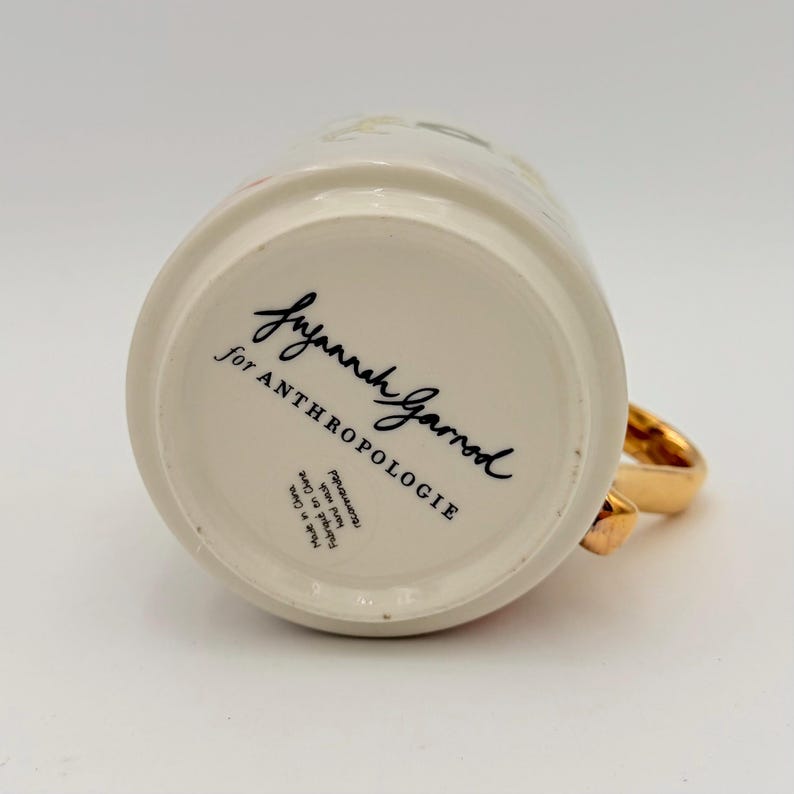 K&ouml;nnte beinhalten: Cremefarbene Keramiktasse mit goldenem Henkel. Auf der Unterseite der Tasse steht in schwarzer Schrift "Suzannah Garrod for ANTHROPOLOGIE". Die Tasse ist in China hergestellt.