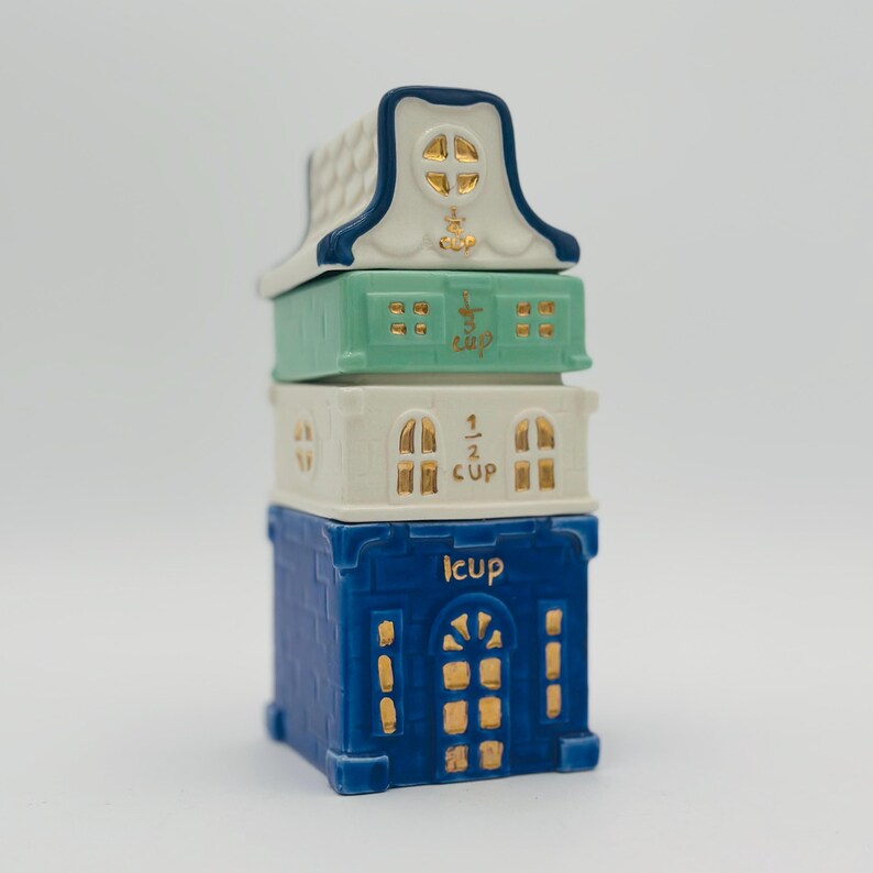 May include: A set of three blue, white, and green ceramic measuring cups shaped like houses. The cups are stacked on top of each other and have gold accents. The top cup says "1/2 cup", the middle cup says "1 cup", and the bottom cup says "1/4 cup".