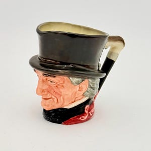 May include: A novelty ceramic mug in the shape of a man's head, complete with a black top hat. The handle is designed as a cane. The face is painted with detail, showing grey hair and a red and black suit.