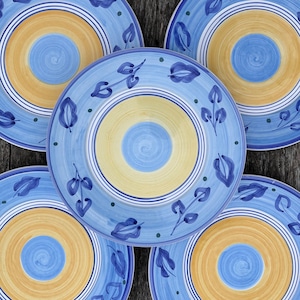 Könnte beinhalten: Vier blaue und gelbe Keramikteller mit einem dekorativen Design. Die Teller haben ein konzentrisches Kreisdesign mit einem blauen Zentrum und einem gelben Außenring. Die Teller sind mit stilisierten Blättern und Punkten verziert.