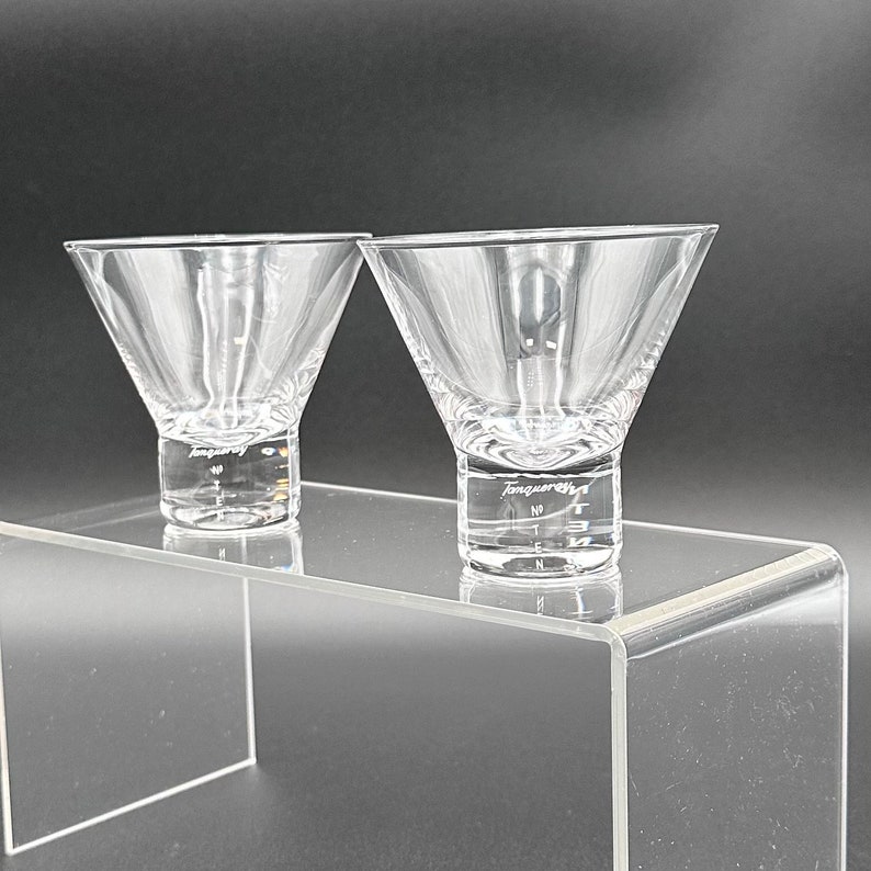 Peut inclure: Deux verres &agrave; martini en verre transparent avec un motif textur&eacute; et le mot "Tangueray" grav&eacute; au fond. Les verres sont pos&eacute;s sur un pr&eacute;sentoir en acrylique transparent.