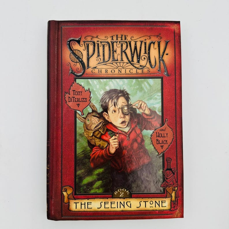 Puede incluir: Un libro de tapa dura titulado "The Spiderwick Chronicles: The Seeing Stone". La portada presenta a un ni&ntilde;o con una lupa, una criatura en su espalda y los nombres de los autores. El libro tiene un borde rojo y una pancarta dorada.