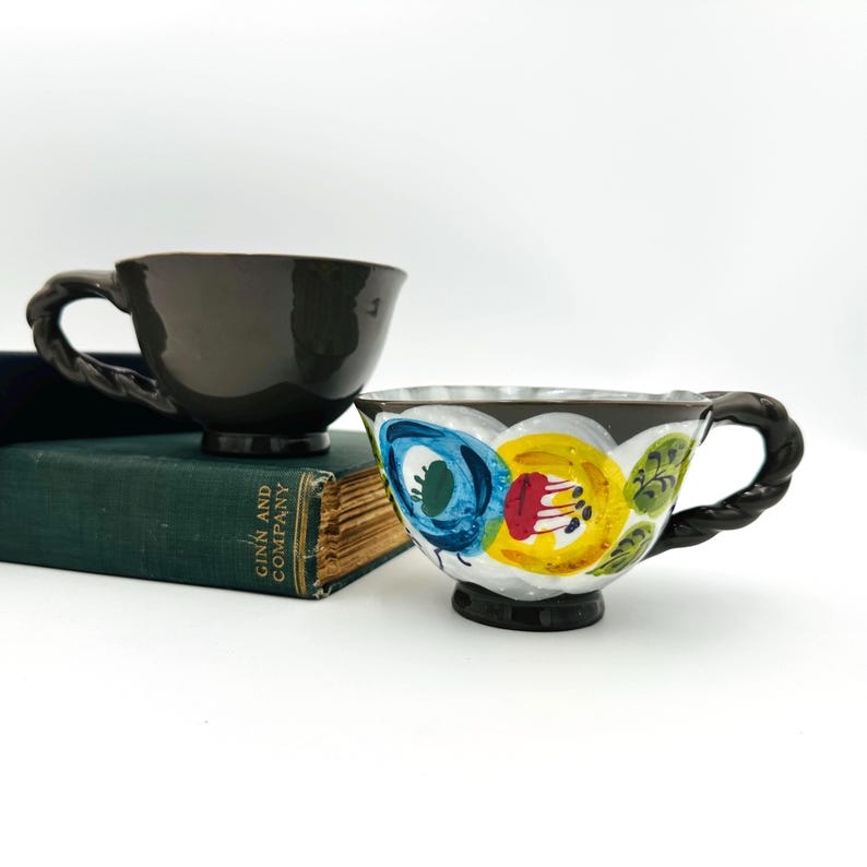 Puede incluir: Dos tazas de t&eacute; de cer&aacute;mica con asas retorcidas. Una es de color marr&oacute;n oscuro liso, la otra es blanca con un dise&ntilde;o floral en azul, amarillo y rojo. Las tazas est&aacute;n sobre un libro verde.