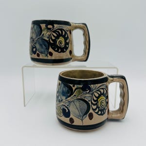 Puede incluir: Dos tazas de cerámica con base beige, borde y asa negros. Las tazas presentan un diseño floral con detalles en azul, verde y amarillo. Las asas son rectangulares.