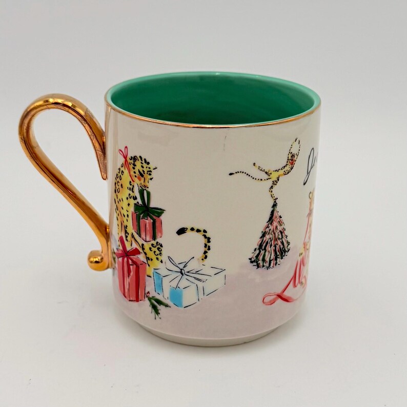 K&ouml;nnte beinhalten: Eine Keramiktasse mit goldenem Henkel und Rand, mit einem skurrilen Design. Die Tasse hat eine cremefarbene Au&szlig;enseite mit Illustrationen eines Leoparden, Geschenken und einer T&auml;nzerin. Der Innenraum ist gr&uuml;n.