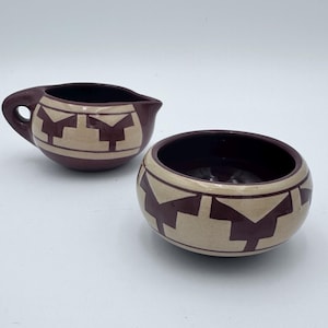 May include: A small, matching creamer and sugar bowl set. The ceramic pieces are a deep burgundy colour with a tan geometric pattern. The creamer has a handle and spout. The sugar bowl is round and open.