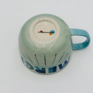 May include: A light blue ceramic mug with a white interior and a blue handle. The mug has a green watercolor design and the word "Wake" written in blue and white on the front. The bottom of the mug has the Anthropologie logo.