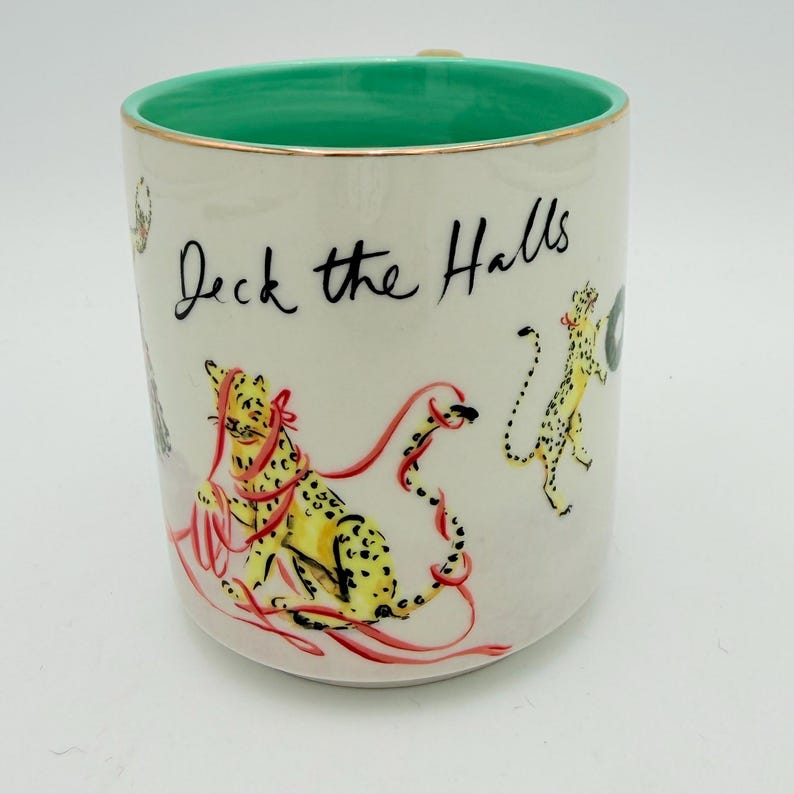 K&ouml;nnte beinhalten: Wei&szlig;er Keramikbecher mit gr&uuml;nem Innenraum und goldenem Rand. Der Becher zeigt den Text "Deck the Halls" in schwarzer Schrift sowie Illustrationen von gelben Geparden und roten B&auml;ndern. Ein festlicher und dekorativer Weihnachtsbecher.