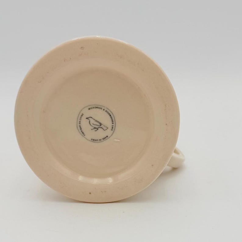 Puede incluir: Una taza de cer&aacute;mica de color crema con una base redondeada y un asa curva. La parte inferior de la taza presenta un logotipo circular con una ilustraci&oacute;n de un p&aacute;jaro y el texto "Microwave & Dishwasher Safe" y "Made in China".