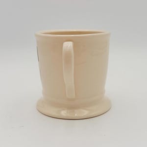 Puede incluir: Una taza de cer&aacute;mica color crema con base redondeada y asa curva. La taza tiene un dise&ntilde;o sencillo y cl&aacute;sico, apto para caf&eacute; o t&eacute;. La taza est&aacute; limpia y parece sin usar.