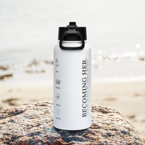 Op de afbeelding: Een witte waterfles met een zwarte dop en handvat. De fles is voorzien van zwarte tekst met de tekst BECOMING HER. NOT BY CHANCE. BY CHOICE. BY GOD. Daaronder staan vier iconen met de labels Pray, Journal, Release en Hydrate.
