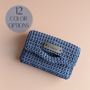 Könnte beinhalten: Eine blaue Häkeltasche mit rechteckiger Klappe und einem grauen Metallschild mit der Aufschrift DV HANDMADE. Eine kreisförmige Grafik in der oberen linken Ecke zeigt den Text 12 COLOR OPTIONS. Die Tasche hat ein strukturiertes Webmuster.