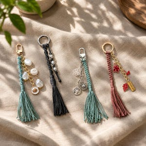 Puede incluir: Cuatro llaveros decorativos con cordones de macramé y borlas. El primero tiene un cordón verde azulado, perlas blancas y un dije de concha. El segundo es negro con cuentas plateadas. El tercero es verde azulado con dijes de árbol y flor. El cuarto es malva con dijes de hojas rojas.