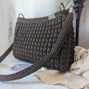 May include: A dark brown, handmade macrame shoulder bag. The bag features a rectangular shape with a braided strap and a small metal tag. The texture of the bag is visible, showcasing the intricate knotting technique. The bag is a stylish accessory.