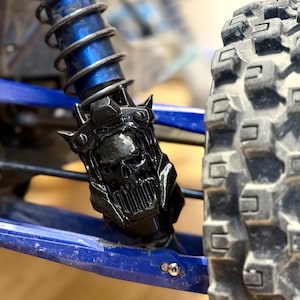 Peut inclure: Gros plan d'un crâne décoratif noir fixé à un système de suspension bleu et argenté. Le crâne présente un design détaillé avec des pointes. Un pneu noir est visible en arrière-plan. Pièce de véhicule.