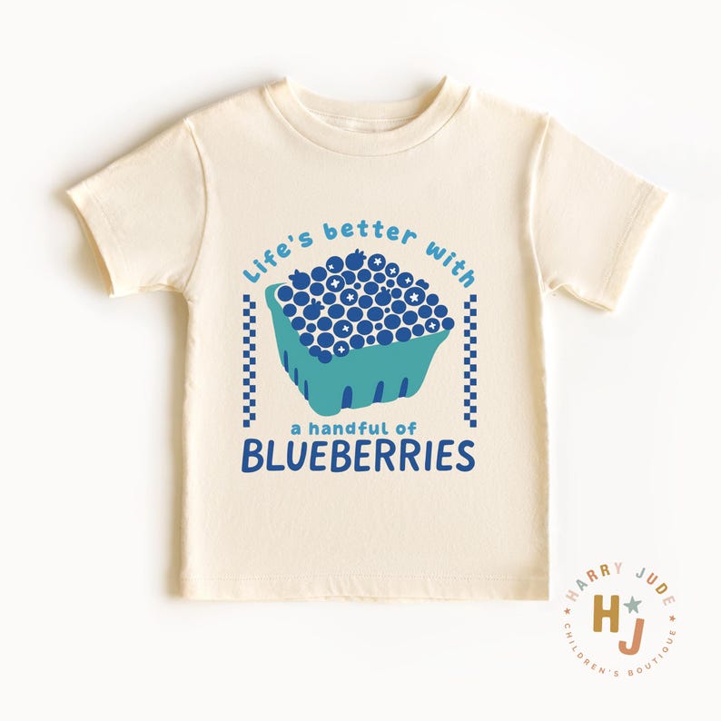 May include: Cream-colored t-shirt featuring a blue and teal graphic of a carton filled with blueberries. The text reads "Life's better with a handful of blueberries."