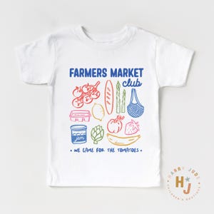 以下が含まれることがあります： カラフルなグラフィックデザインの白いTシャツ。デザインには「FARMERS MARKET club」の文字と「WE CAME FOR THE TOMATOES」のフレーズが含まれています。トマト、バゲット、イチゴなど、さまざまな果物や野菜のイラストも含まれています。