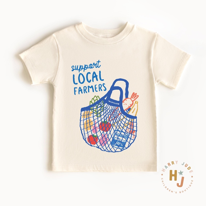 Op de afbeelding: Cr&egrave;mekleurig T-shirt met een blauwe afbeelding van een tas vol groenten en fruit. De tekst "support LOCAL FARMERS" staat in blauw boven de tas. Het shirt heeft korte mouwen en een ronde hals. Een klein logo staat rechtsonder.