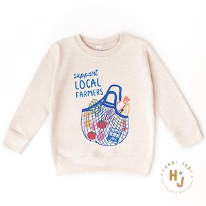 Op de afbeelding: Een beige sweatshirt met een blauwe afbeelding van een herbruikbare boodschappentas gevuld met producten. De tekst "support LOCAL FARMERS" staat in blauw boven de tas. De sweatshirt heeft lange mouwen en een ronde hals.