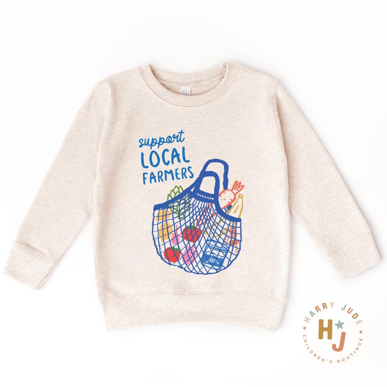 Op de afbeelding: Een beige sweatshirt met een blauwe afbeelding van een herbruikbare boodschappentas gevuld met producten. De tekst "support LOCAL FARMERS" staat in blauw boven de tas. De sweatshirt heeft lange mouwen en een ronde hals.