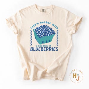 May include: Cream-colored t-shirt with a blue and teal graphic of blueberries in a basket. The text reads "Life's better with a handful of blueberries."