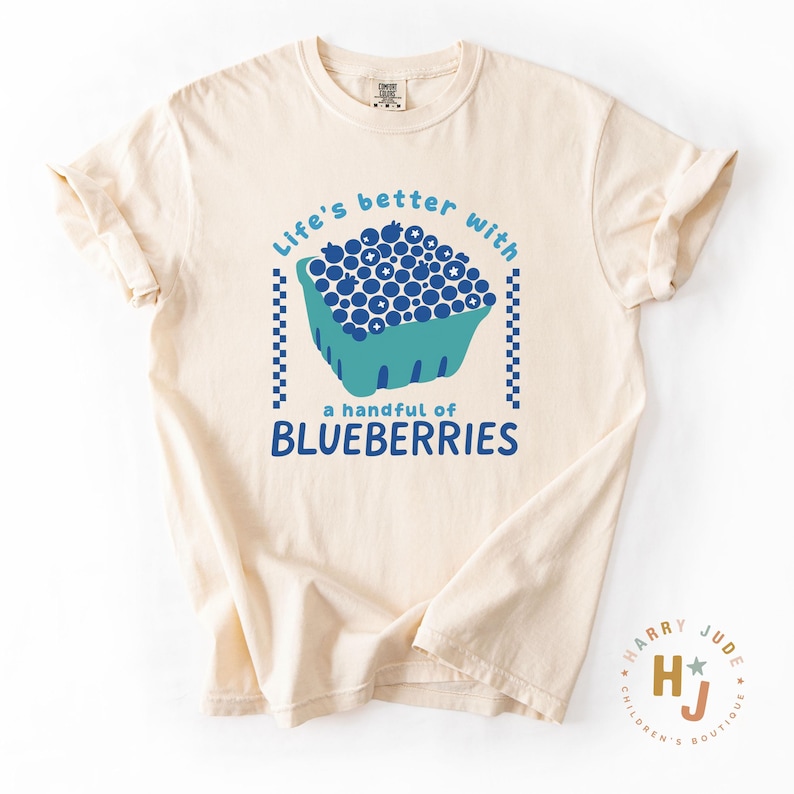 May include: Cream-colored t-shirt with a blue and teal graphic of blueberries in a basket. The text reads "Life's better with a handful of blueberries."