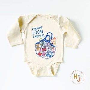 Op de afbeelding: Cr&egrave;me babybody met lange mouwen. De voorkant heeft een blauwe afbeelding van een herbruikbare boodschappentas gevuld met producten, en de tekst "support LOCAL FARMERS" in blauw. Een klein logo staat in de rechter benedenhoek.