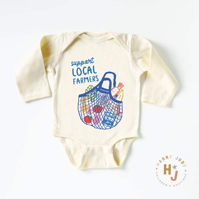 Op de afbeelding: Cr&egrave;me babybody met lange mouwen. De voorkant heeft een blauwe afbeelding van een herbruikbare boodschappentas gevuld met producten, en de tekst "support LOCAL FARMERS" in blauw. Een klein logo staat in de rechter benedenhoek.