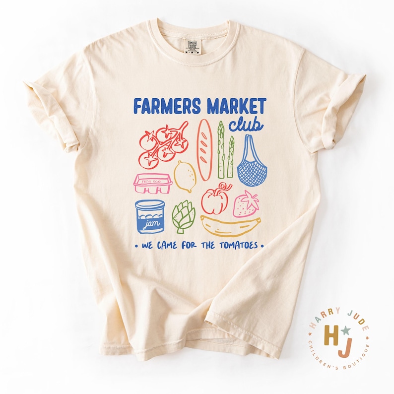 以下が含まれることがあります： クリーム色のTシャツ。カラフルなファーマーズマーケットのイラストと、青色の「FARMERS MARKET club」の文字がプリントされています。イラストの下には「WE CAME FOR THE TOMATOES」の文字。右下にはロゴがあります。