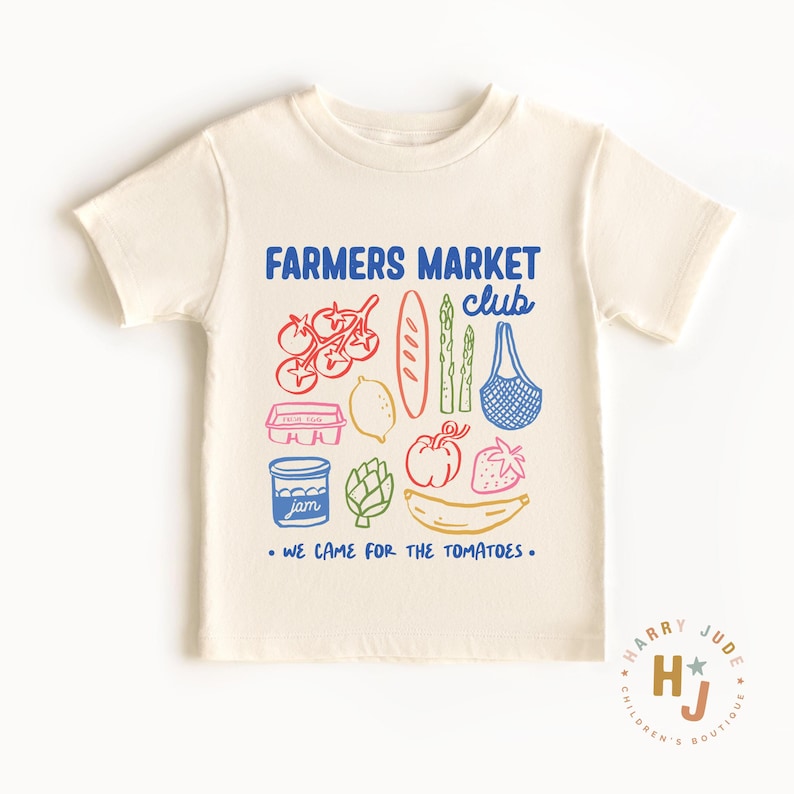 以下が含まれることがあります： クリーム色のTシャツ。カラフルな農産物のグラフィックと「FARMERS MARKET CLUB」の文字が特徴です。下には「WE CAME FOR THE TOMATOES」と書かれています。デザインにはトマト、パン、アスパラガス、買い物袋が含まれています。