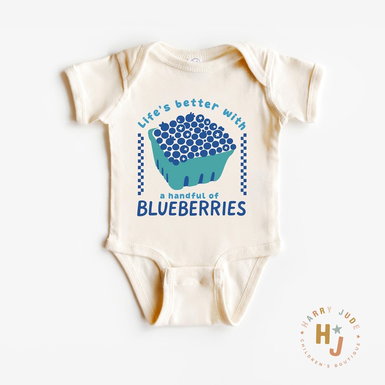 May include: Cream-colored baby onesie with a blue and teal graphic of a blueberry basket and the text "Life's better with a handful of BLUEBERRIES." The onesie has short sleeves and a round neckline. The brand logo is in the bottom right corner.