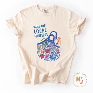 Op de afbeelding: Cr&egrave;mekleurig T-shirt met de blauwe tekst "support LOCAL FARMERS". Een blauwe nettas-afbeelding is gevuld met kleurrijke producten. Het shirt heeft korte mouwen en een ronde hals.