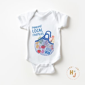 Op de afbeelding: Witte babybody met een blauwe afbeelding van een boodschappentas gevuld met producten. De tekst "support LOCAL FARMERS" staat boven de tas. De body heeft korte mouwen en een drukknoopsluiting.