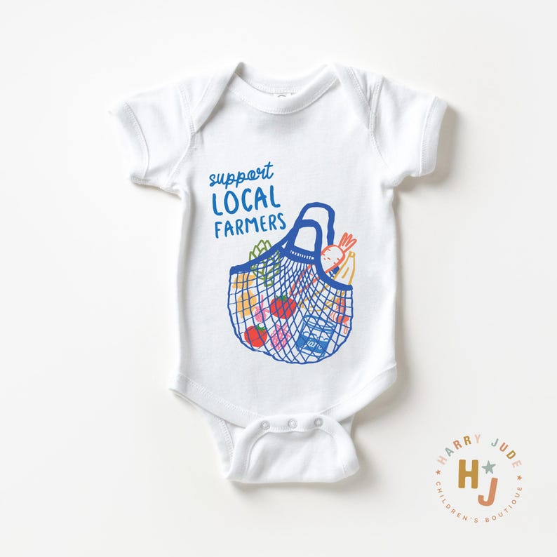 Op de afbeelding: Witte babybody met een blauwe afbeelding van een boodschappentas gevuld met producten. De tekst "support LOCAL FARMERS" staat boven de tas. De body heeft korte mouwen en een drukknoopsluiting.