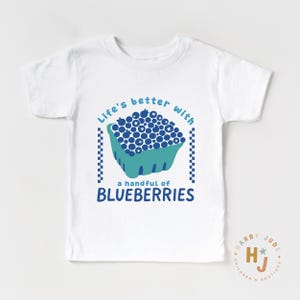 May include: White t-shirt with a blue and teal graphic. The graphic features a carton of blueberries and the text "Life's better with a handful of BLUEBERRIES". The shirt has short sleeves and a crew neck.