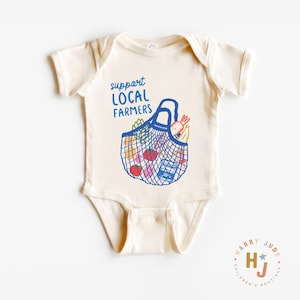 Op de afbeelding: Cr&egrave;me babyromper met de blauwe tekst "support LOCAL FARMERS". Een blauwe nettas-afbeelding is gevuld met kleurrijke producten. De romper is een kledingstuk uit &eacute;&eacute;n stuk met korte mouwen, voor baby's en peuters.