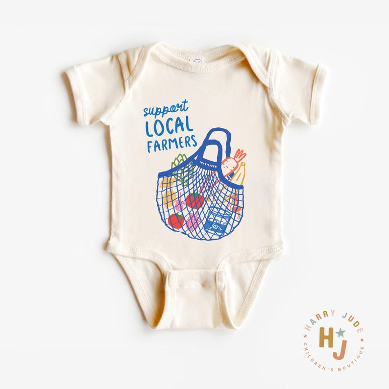 Op de afbeelding: Cr&egrave;me babyromper met de blauwe tekst "support LOCAL FARMERS". Een blauwe nettas-afbeelding is gevuld met kleurrijke producten. De romper is een kledingstuk uit &eacute;&eacute;n stuk met korte mouwen, voor baby's en peuters.