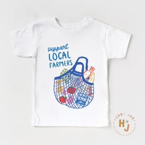 Op de afbeelding: Wit T-shirt met een blauwe illustratie van een boodschappentas vol producten. De tekst "support LOCAL FARMERS" staat boven de tas. Het shirt heeft korte mouwen en een ronde hals.