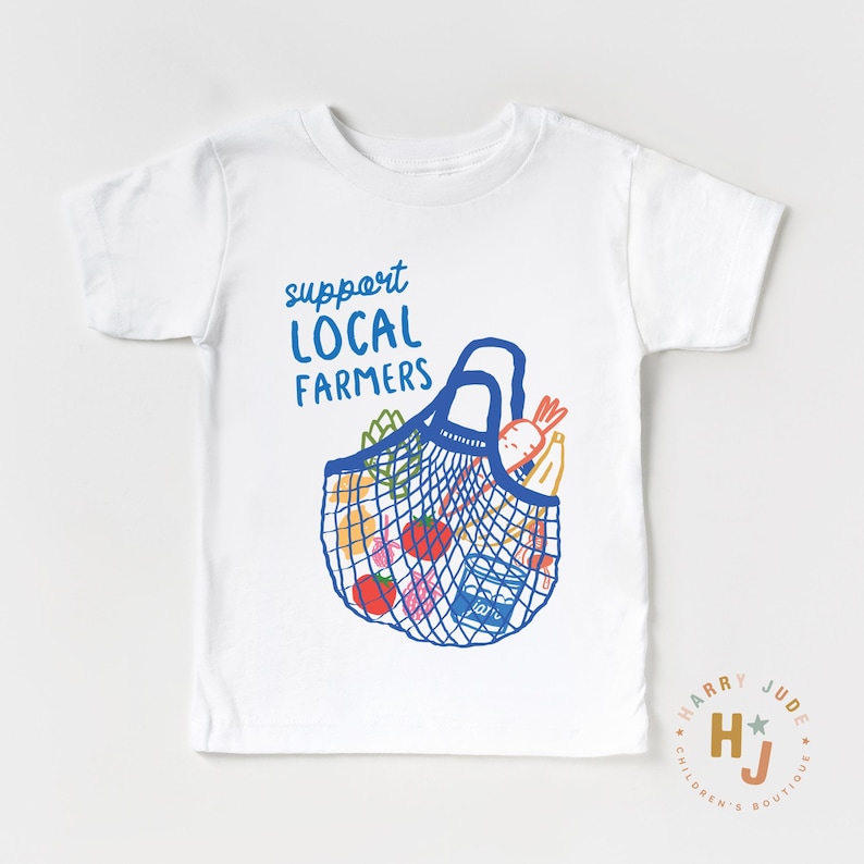 Op de afbeelding: Wit T-shirt met een blauwe illustratie van een boodschappentas vol producten. De tekst "support LOCAL FARMERS" staat boven de tas. Het shirt heeft korte mouwen en een ronde hals.