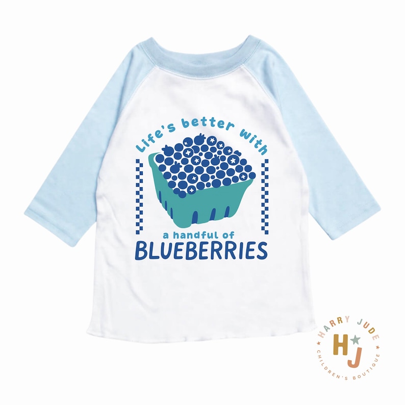 May include: A white and light blue raglan-style shirt with a graphic of blueberries in a basket. The text on the shirt reads "Life's better with a handful of blueberries."