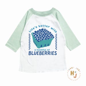 May include: White and light green raglan-sleeve shirt with the text "Life's better with a handful of BLUEBERRIES" and a graphic of a carton of blueberries. The shirt has a round neck and three-quarter length sleeves.