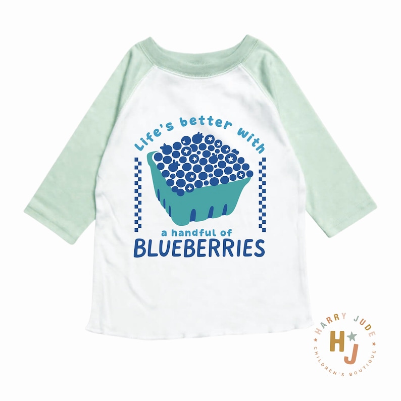 May include: White and light green raglan-sleeve shirt with the text "Life's better with a handful of BLUEBERRIES" and a graphic of a carton of blueberries. The shirt has a round neck and three-quarter length sleeves.