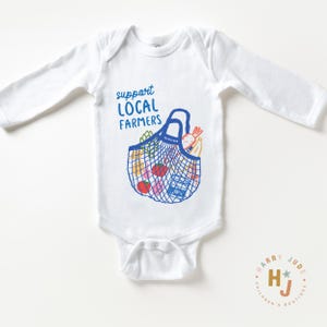 Op de afbeelding: Witte babybody met lange mouwen. Het heeft een blauwe afbeelding van een boodschappentas vol producten en de tekst "support LOCAL FARMERS" in het blauw. Een logo voor "Harry Jude Children's Boutique" staat rechtsonder.