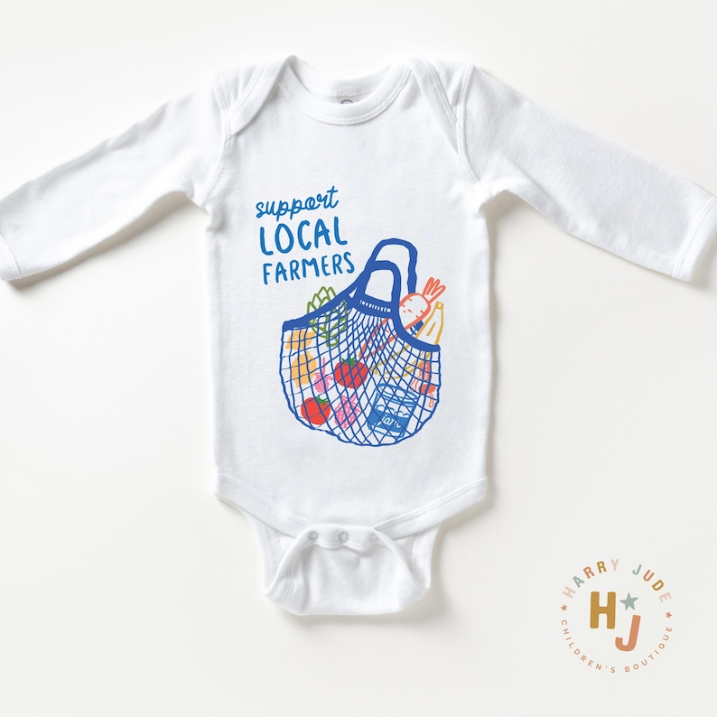 Op de afbeelding: Witte babybody met lange mouwen. Het heeft een blauwe afbeelding van een boodschappentas vol producten en de tekst "support LOCAL FARMERS" in het blauw. Een logo voor "Harry Jude Children's Boutique" staat rechtsonder.