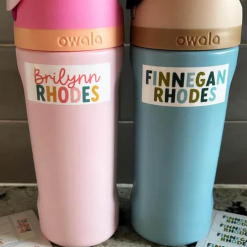 May include: Two Owala water bottles, one pink and one blue, with colorful lids. Each bottle has a personalized label. The pink bottle reads "Brilynn RHODES" and the blue bottle reads "FINNEGAN RHODES".